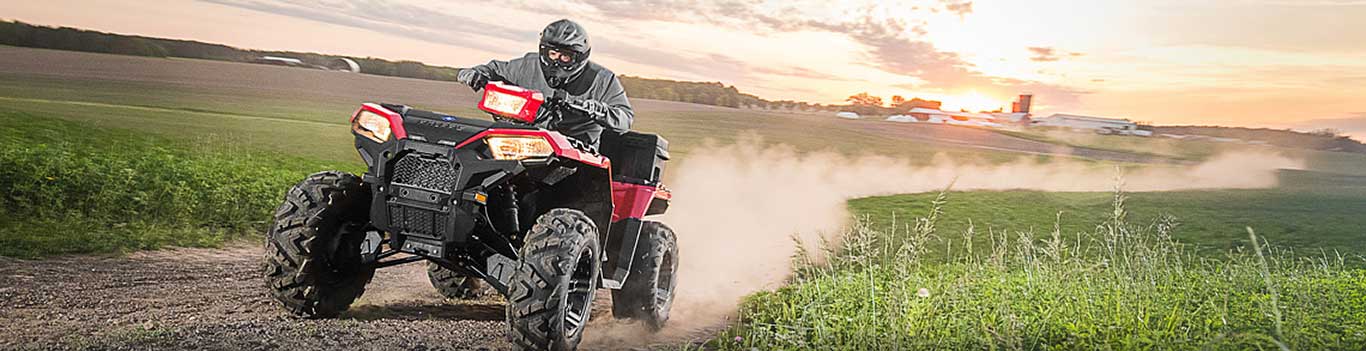 Man rides a red ATV across a dirt path near farm fields.